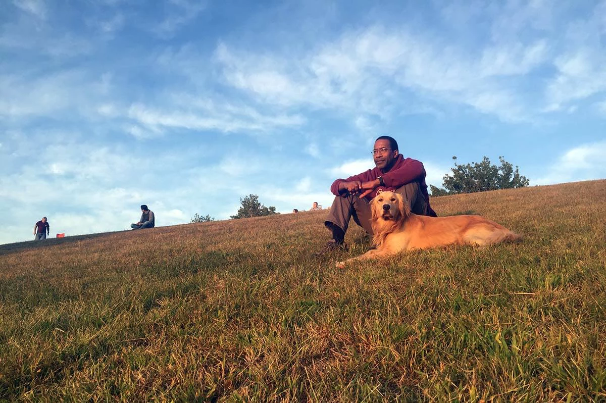Shawn Hines sitting down in a grass field with a Golden Retriever lying next to him.